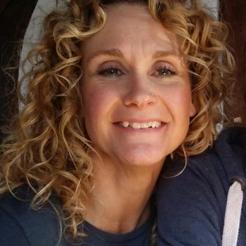 White female with curly blonde hair. Blue eyes and smiling into the camera, wearing a navy blue round neck top.