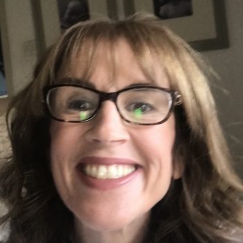 White female with shoulder length brown, wavy hair. Smiling at the camera, wearing a white jumper and black framed glasses.