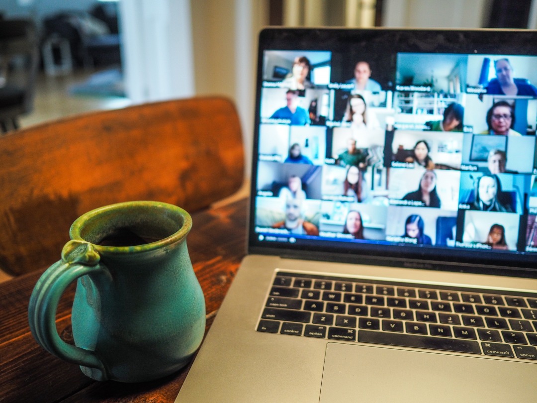 Silver Mac laptop with black button. Green tea or coffee cup sat next to it on a brown desk. Screen on laptop shows a Zoom meeting with 16 people around the screen