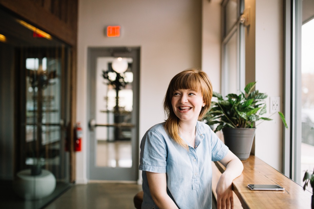 Female standing in a bright airy room. She is leaning on a window shelf, smiling, with light, long hair, fringe. Wearing a grey t-shirt