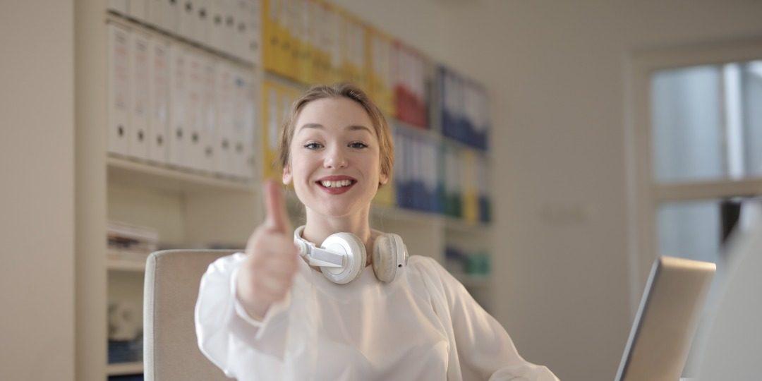 Girl in white oversized blouse, white headphones around her neck. She is giving the thumbs up and sitting at a desk with a laptop.