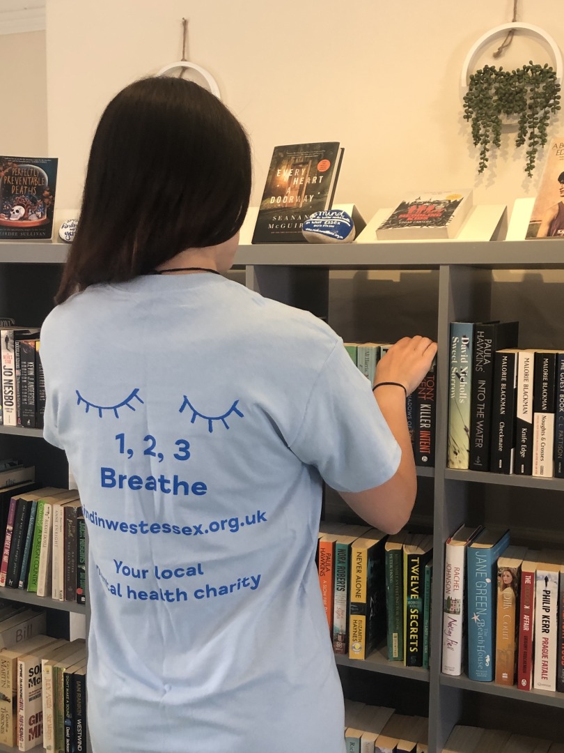 Female, with long dark hair standing with her back to the camera looking through a bookcase. On the light blue tee-shirt is 2 closed eyelids with the text Breathe 1,2,3, website address of MindinWestEssex.org.uk and the text ‘Your local mental health charity'