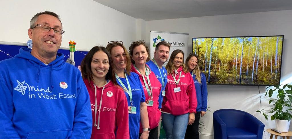 7 people wearing pink and blue branded hoodies. Stood in a row in a white room with a TV mounted on the wall.