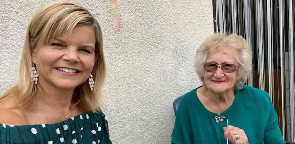 Two women sat aside both wearing teal green tops, one plain and one with spots. Eating dessert and smiling at the camera. One is an older women wearing sun glasses and the other is younger with shoulder length blonde hair.