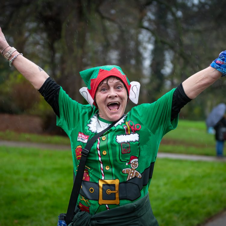 A woman in an elf hat and t-shirt raising both arms as she looks at the camera during the run